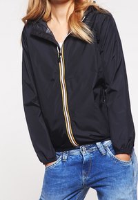 Svart lättviktsjacka med huva, som har en vit och gul dragkedja, elastiska ärmslut och en avslappnad passform, matchad med blå denimjeans.
