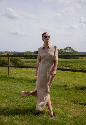 Kobieta w beżowej sukience na guziki oraz okularach przeciwsłonecznych, idąca po trawie, w tle drewniane ogrodzenie i zielone pola.