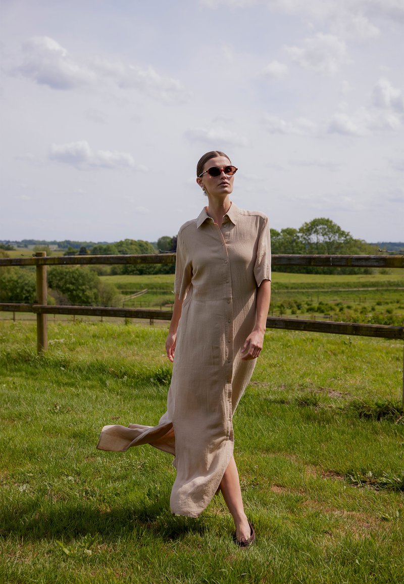 Vrouw die een beige jurk met knopen en een zonnebril draagt, loopt op gras met een houten hek en groene velden op de achtergrond.
