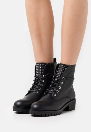Bottines noires en matériau texturé, avec un laçage à l'avant, des clous argentés sur le col et une semelle épaisse et à motifs.
