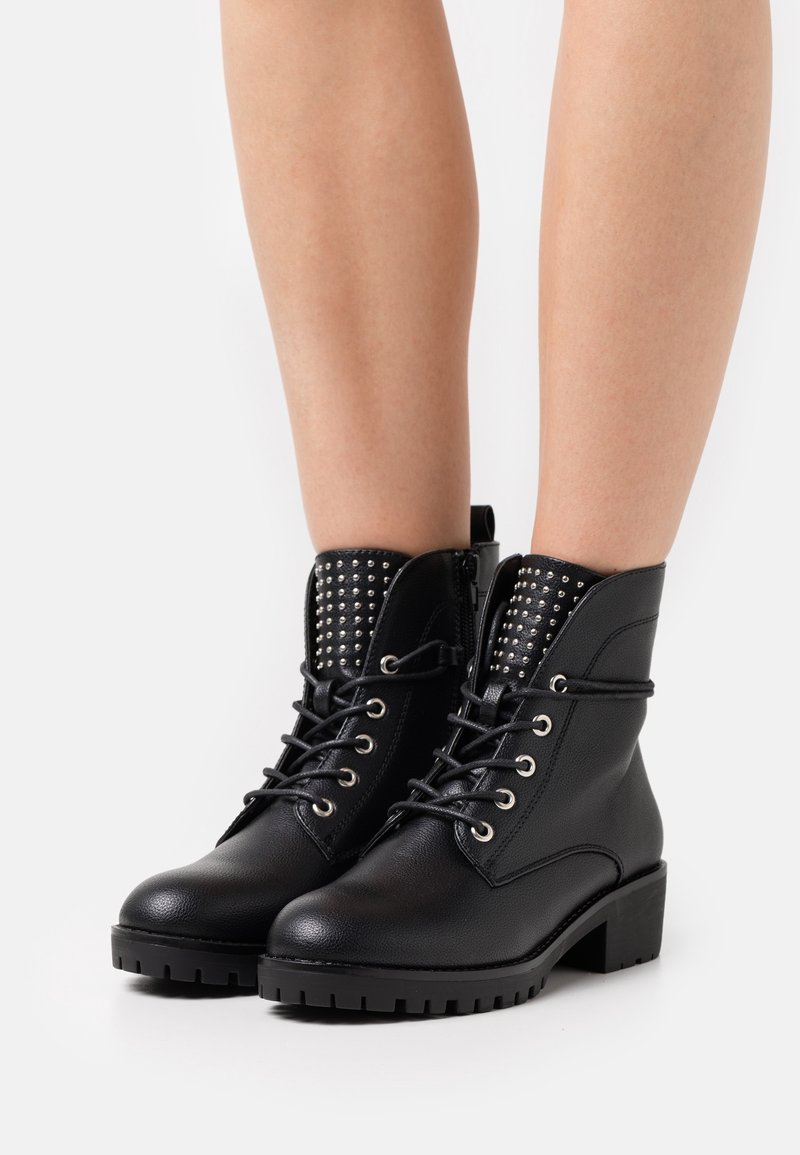 Botines negros de tobillo hechos de material texturizado, con un frente de cordones, tachuelas plateadas en el cuello y una suela gruesa y con patrón.