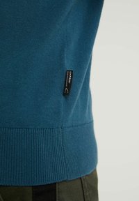 Pull en tricot bleu sur le torse supérieur montrant un ourlet côtelé et une petite étiquette de marque noire indiquant "CHASIN' Denim Craftsmanship".