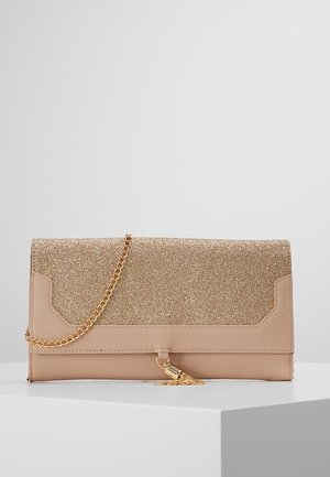 Beigen värinen clutch-laukku, jossa kimaltava kultainen läppä, kultainen ketjuhihna ja kultainen tupsukoriste, asetettuna valkoiselle jalustalle yksiväristä taustaa vasten.