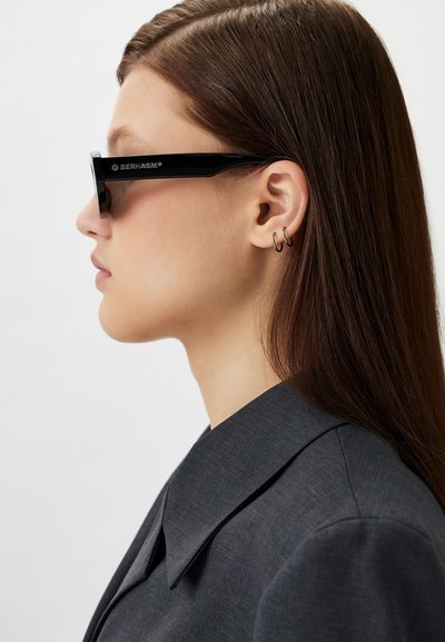 Black rectangular sunglasses with thick frames. The lenses are dark, and the side arm features a logo. Worn with a dark collared blazer.