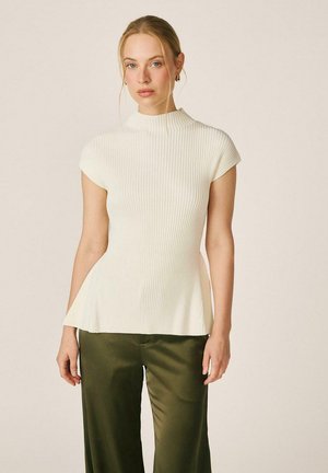 Donna con capelli biondi indossa una maglietta bianca a coste con maniche corte e pantaloni verde oliva a gamba larga su uno sfondo beige uniforme.
