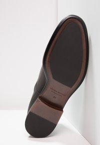 Chaussure en cuir noir à bout arrondi, avec une surface lisse et une semelle marron texturée. Comprend le logo "VAGABOND" et la taille 42.