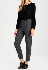 Top nero con zip frontale, abbinato a pantaloni su misura grigi a quadri con risvolti e scarpe stringate nere.