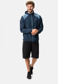 Marineblaue wasserdichte Jacke mit hellblauen Schulterakzenten, durchgehendem Reißverschluss und Kapuze. Kombiniert mit schwarzen Shorts und schwarzen Schuhen.