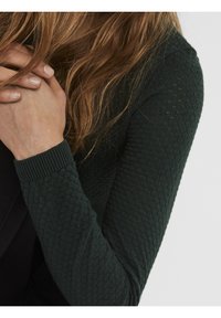 Personne aux longs cheveux bruns portant un pull vert foncé texturé, les mains jointes près du menton, sur un fond uni.