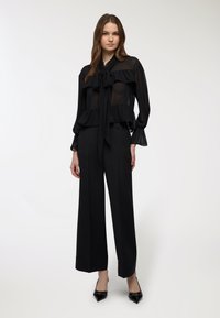 Blusa nera trasparente con un fiocco al collo, dettagli arricciati e maniche lunghe, abbinata a pantaloni neri a vita alta e gamba larga, e tacchi.