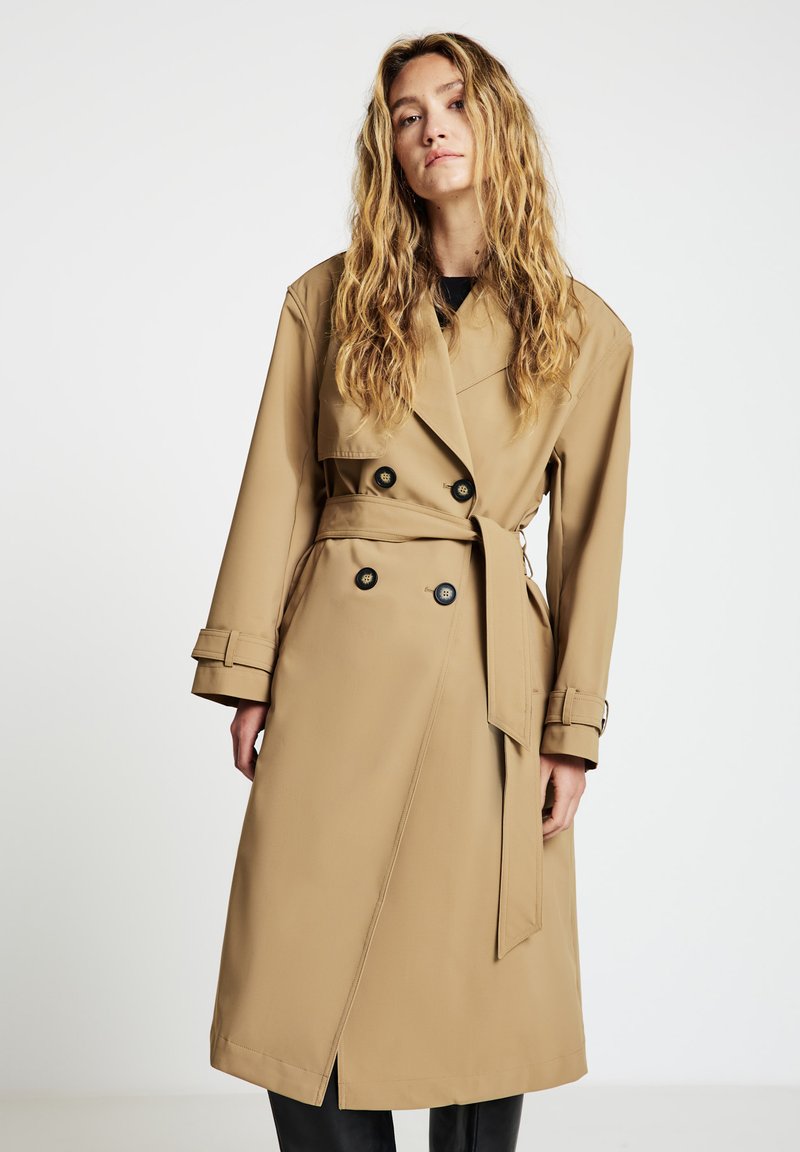 Beiger Trenchcoat mit doppelt geknöpftem Vorderteil, schwarzen Knöpfen, tailliertem Gürtel und weitem Kragen. Hergestellt aus einem glatten Stoff mit geradem Schnitt.