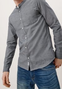 Grijs overhemd met knopen en een mandarijnkraag, voorzien van een borstzak, textuurstof en witte knopen. Gecombineerd met een blauwe spijkerbroek.