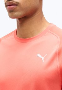 Κοντινό πλάνο ενός ατόμου που φορά ένα κοραλλί αθλητικό πουκάμισο με λευκό λογότυπο Puma στο στήθος, πάνω σε απλό φόντο.