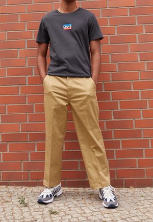 Personne debout contre un mur en briques rouges, portant un T-shirt gris foncé avec un logo, un pantalon beige et des baskets blanches et grises, les mains dans les poches.