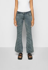 Jeans flare em denim azul desbotado com padrões de costura diagonal, cintura alta, bolsos frontais e fecho de botão. Combinados com sandálias pretas de plataforma.