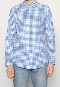 Blå och vit rutigt skjorta med knappstängning, långa ärmar, krage och en liten logotyp på bröstet. Tyget verkar slätt med ett rutmönster.