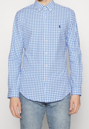 Camicia a maniche lunghe a quadri blu e bianchi con colletto e un piccolo logo sul petto. Il tessuto sembra liscio con un motivo a scacchi.