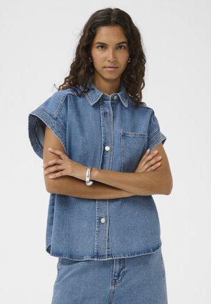 Žena s kučeravými vlasmi, oblečená v bezrukávovej rifľovej košeli na gombíky a rifliach, so založenými rukami a neutrálne výrazom, na jednoduchom bielom pozadí.