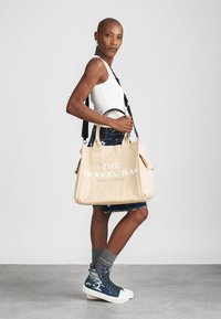 Bež platnena torba s natpisom "TORBA ZA PUTOVANJE", dvostrukim gornjim ručkama i podesivim remenom za rame. Ima bočne džepove i strukturirani oblik.