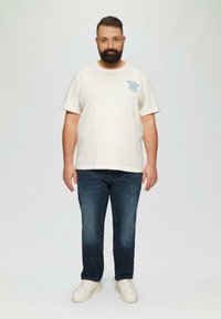 Lichtbeige katoenen T-shirt met blauwe bedrukte tekst op de linker borst, gecombineerd met donkerblauwe jeans en witte sneakers. Eenvoudig, casual ontwerp.