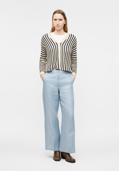 Žena stojaca s rukami v kapsách svetlomodrých nohavíc s širokými nohavicami, oblečená v čierno-bielom pruhovanom kardigane cez biele tričko a hnedé topánky.