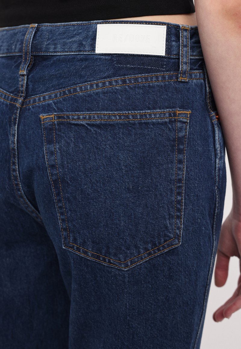 Dunkelblaue Jeans aus Denim verfügen über ein weißes Herstelleretikett am Bund, orangefarbene Nähte und Gesäßtaschen mit einer dezenten, strukturierten Oberfläche.