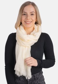 Écharpe crème en tissu léger et texturé avec des bords effilochés, drapée autour du cou, tenue par une personne portant un haut noir.