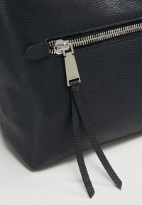 Gros plan d’un sac en cuir noir texturé avec une fermeture éclair argentée et deux longues tirettes en cuir.