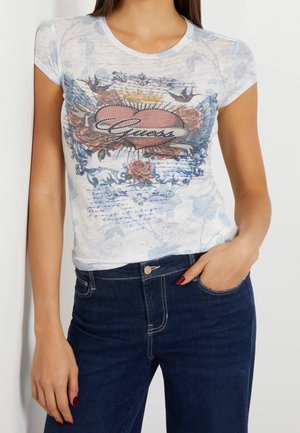 Frau trägt ein weißes, figurbetontes T-Shirt mit Herz-, Vogel- und Blumenmotiv sowie "Guess"-Schriftzug, kombiniert mit dunkelblauen Jeans.