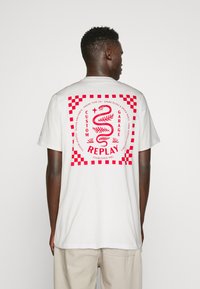 Camiseta de algodón blanca con un gran gráfico rojo en la espalda. Presenta un diseño de serpiente, bordes a cuadros y un texto que dice "REPETICIÓN DEL GARAGE PERSONALIZADO."