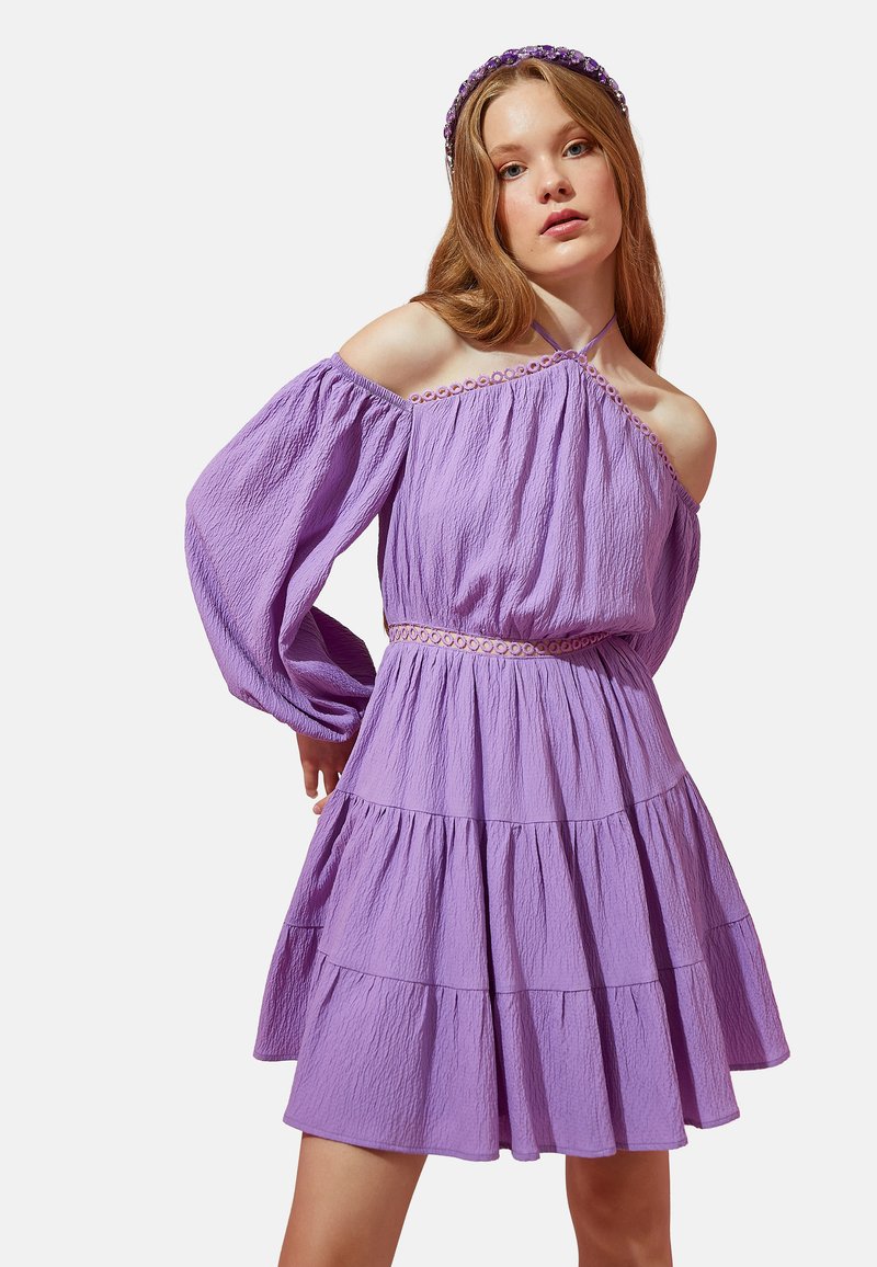 Lila Kleid mit schulterfreien Ärmeln, strukturiertem Stoff, gestuften Rüschendetails und tailliertem Schnitt. Die Accessoires umfassen ein passendes Haarband.