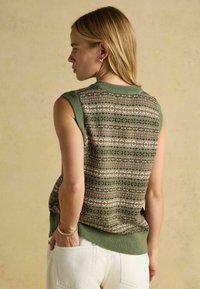 Blond naine, kes kannab ilma varrukat roheline mustriga kudumvest ja valgeid pükse, seisab külje peal, üks käsi taskus, beežil taustal.