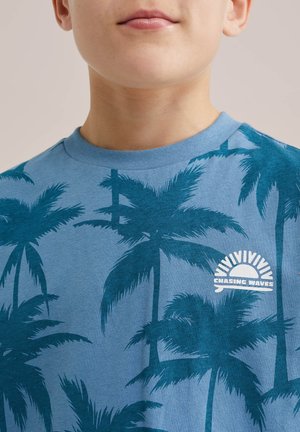 Kind draagt een blauw t-shirt met een donker teal palmbomenpatroon en een wit "chasing waves" zonlogo op de borst tegen een effen achtergrond.