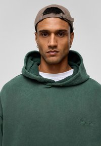 Overmaatse groene hoodie met een textuurrijke capuchon, voorzien van een klein logo detail. Een licht t-shirt is zichtbaar eronder. Een bruine pet voegt een casual accent toe.