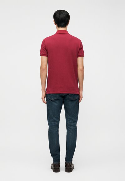 Άνδρας που στέκεται με την πλάτη προς τα εμπρός, φορώντας κόκκινο πόλο με κοντά μανίκια, σκούρο μπλε τζιν και καφέ παπούτσια σε λευκό φόντο.