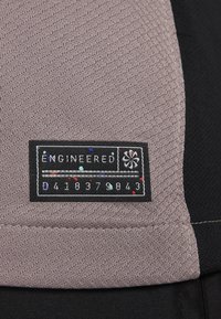 Etiqueta de prenda con el texto "ENGINEERED" en blanco sobre un fondo negro, cosida en una tela texturizada color topo con acentos coloridos.