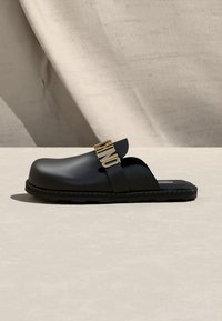 Sabot noir à enfiler avec une semelle épaisse et une lanière dorée "MOSCHINO" sur le dessus, présenté sur un fond beige texturé.