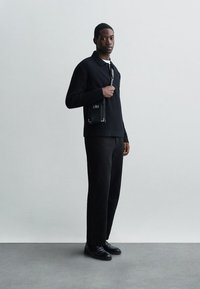 Moški v črnem rebrastem puloverju, črnih hlačah in črnih čevljih, drži majhno črno torbico čez ramo, stoji ob enakomernem svetlo sivo steni.