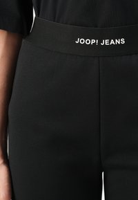 Melni elastīgi bikses ar platu jostu, kurā redzams "JOOP! JEANS" logo baltā krāsā. Gluda tekstūra, pielāgota formas, un minimālais dizains.