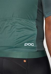 Zelený cyklistický dres s texturovanou tkaninou, obsahující boční kapsu, elastický lem a bílé logo "poc". Černé cyklistické šortky jsou viditelné níže.