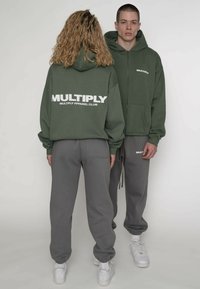 Grøn hættetrøje med hvid "MULTIPLY" tekst, kombineret med grå joggingbukser med matchende logo, komplet med hvide sneakers.