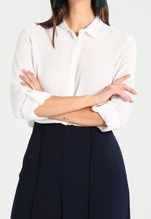 Person with arms crossed wearing a white buttoned shirt with rolled sleeves and high-waisted black pants, showing a small wrist tattoo.