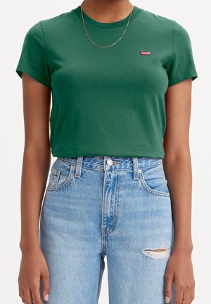 Personne portant un t-shirt vert Levi's, un collier chaîne doré et un jean taille haute bleu clair avec une petite déchirure sur la cuisse droite.