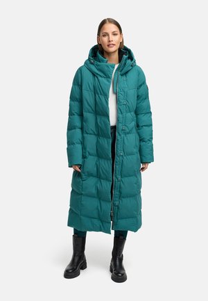 Türkisfarbener Puffer-Mantel mit Kapuze, der ein gestepptes Design und Druckknöpfe aufweist, getragen über einem weißen Hemd und dunklen Jeans, kombiniert mit schwarzen Stiefeln.