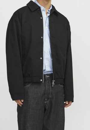 Homme portant une veste bomber noire sur une chemise rayée bleu clair à boutons et un jean bleu foncé avec des coutures visibles.