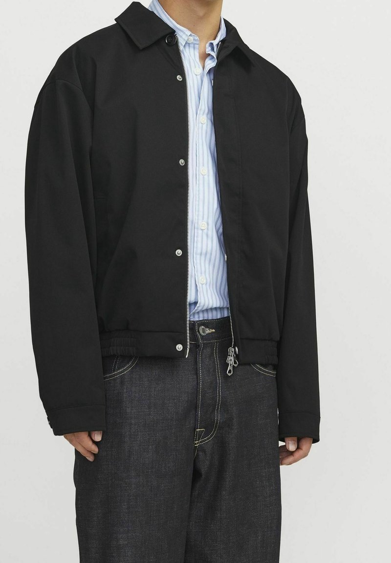 Homme portant une veste bomber noire sur une chemise rayée bleu clair à boutons et un jean bleu foncé avec des coutures visibles.
