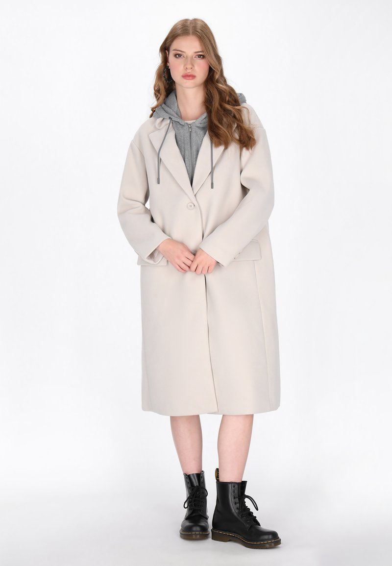 Cappotto beige in lana con colletto a contrasto e chiusura frontale con bottoni, indossato sopra una felpa con cappuccio grigia. Stivaloni neri con lacci completano il look.
