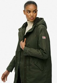 Parka olive en tissu durable, dotée d'une capuche, d'une taille réglable par cordon et d'un patch avec un drapeau américain sur la manche.