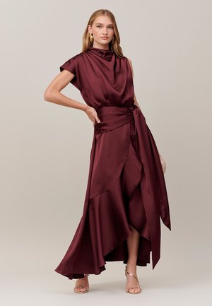 Femme portant une robe en satin rouge foncé sans manches avec un corsage drapé et une jupe fluide asymétrique, associée à des boucles d'oreilles dorées et des talons à brides.