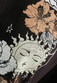 Mustassa kankaassa on painettuja kuvioita, joissa on rauhallisen kasvojen omaava aurinko, jota ympäröi beige- ja oranssipainotteisia kukkia sekä valkoisia tähtiä.